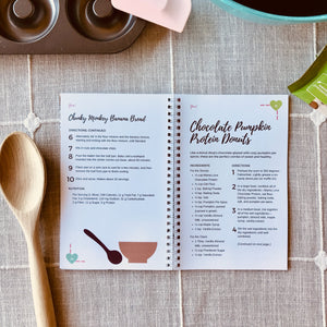 "Yum! Protein-Packed Smoothies & Snacks for Chocolate Lovers" Cookbook, open to page 34, on a surface with a tablecloth, mixing spoons, and other baking tools