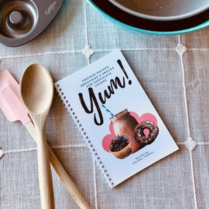 "Yum! Protein-Packed Smoothies & Snacks for Chocolate Lovers" Cookbook on a surface with a tablecloth, mixing spoons, and other baking tools